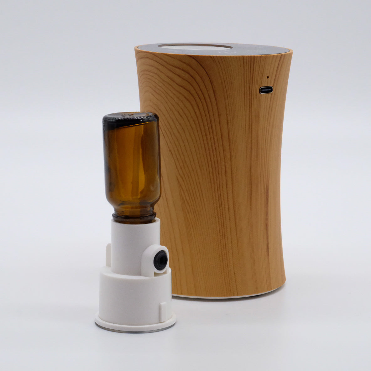 Nébulisateur avec adaptateur blanc et bouteille ambrée vissée tête en bas; base finition bois avec port USB-C en arrière-plan.